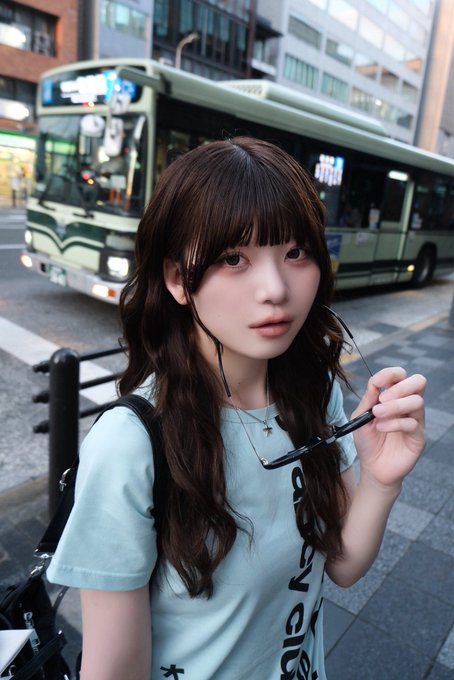 A young woman with long brown hair stands on a city street holding her eyeglasses, with a green public bus visible in the background.