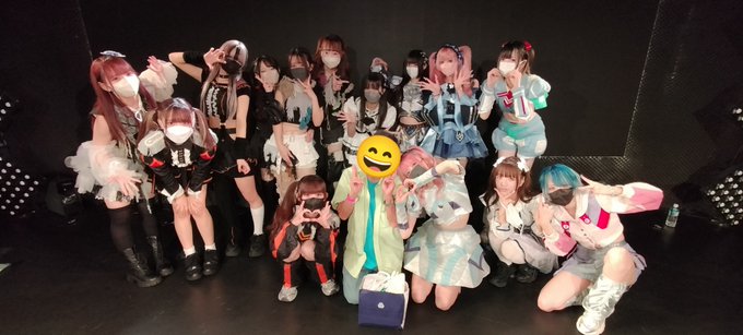 A group photo of people wearing various colorful, elaborate costumes, many with face masks, posing together; one person's face is covered by a yellow smiley face emoji.