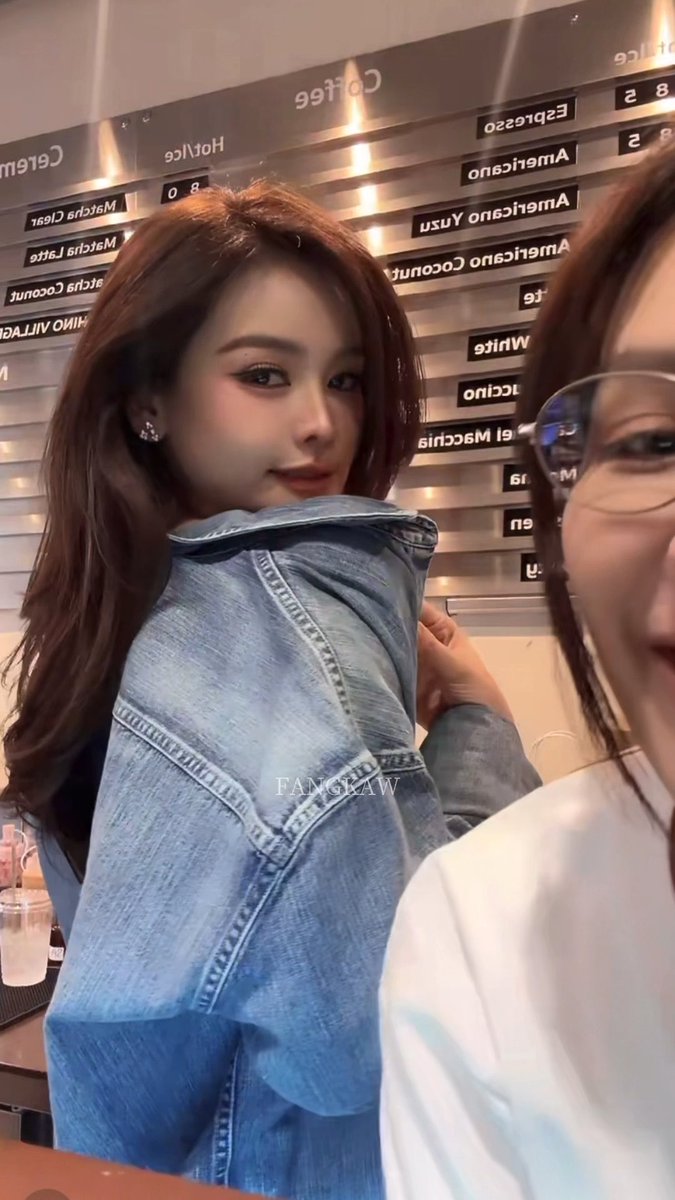 A young woman with long brown hair, wearing a denim jacket and earrings, poses for a photo. The background appears to be a cafe menu board, and the watermark "FANIGLAW" is visible across the image.