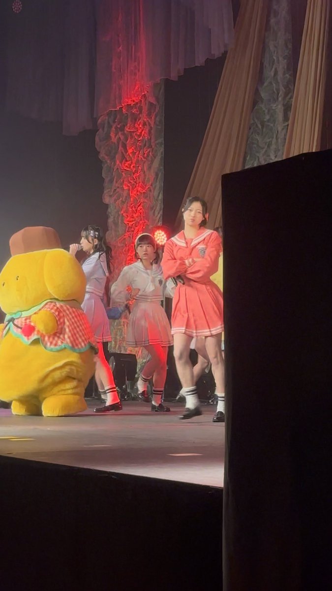 Three performers in pink and white sailor outfits standing near a yellow bear mascot on a stage with dramatic red lighting and fabric backdrop.