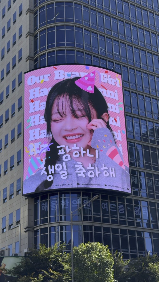 A large outdoor digital billboard on a glass high-rise displays a pink birthday advertisement for 'Haewon,' featuring her face, a party hat, and Korean text wishing a happy birthday.