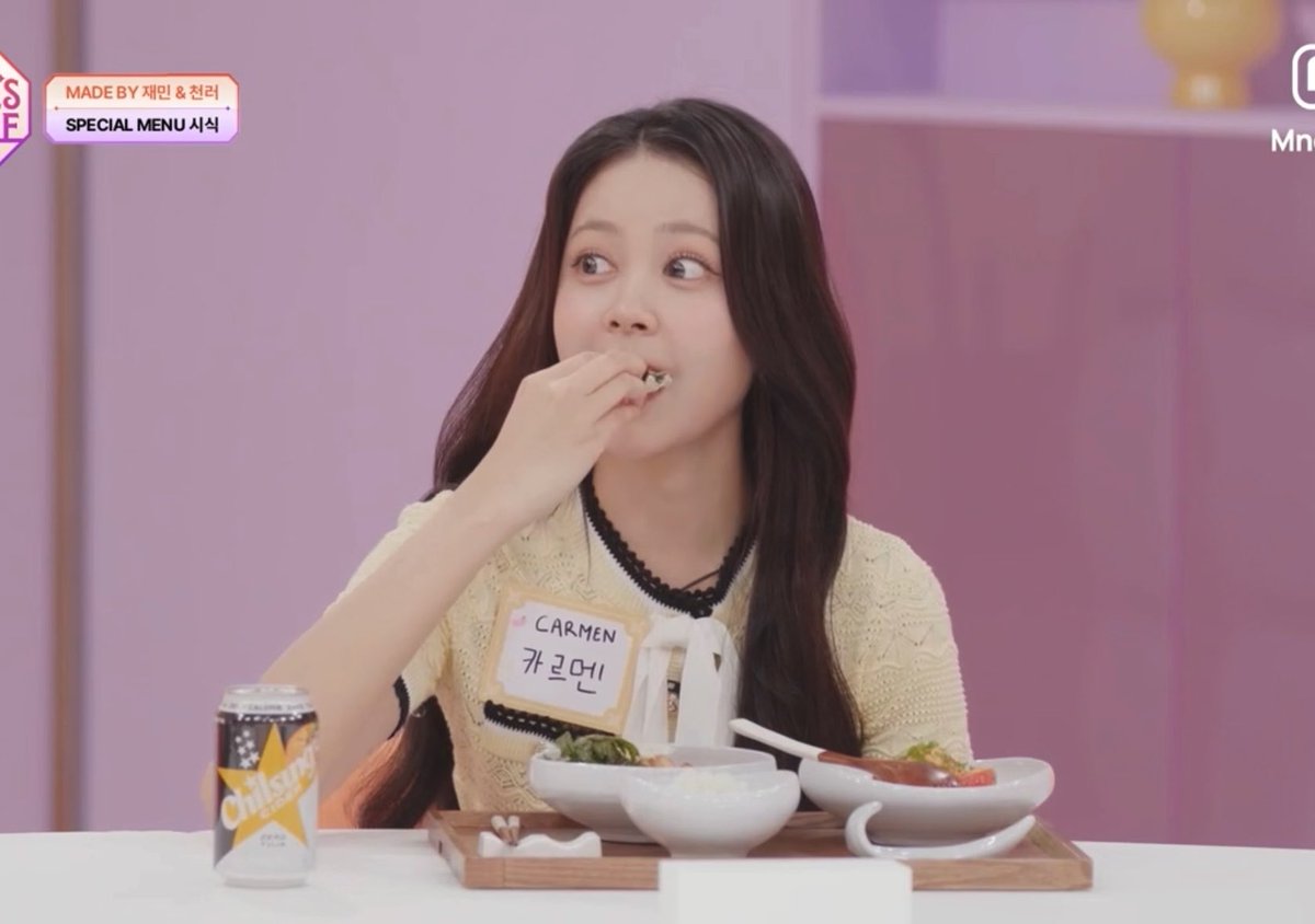 A young woman wearing a yellow top and a name tag reading "CARMEN" eats a small piece of food at a table with a Carlsberg beer, rice, and side dishes, during an "IDOL'S TABLE" broadcast marked "SPECIAL MENU" by J-Min and Hyeong-gyun. Her bright, charming smile as she enjoys the seaweed complements the post's praise of her cuteness and delightful personality.