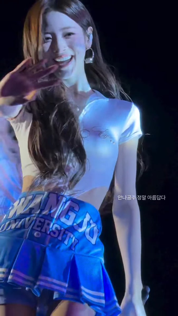A smiling woman with long dark hair waves toward the camera while wearing a light blue crop top and a blue "EWHA UNIVERSITY" cheerleader skirt, with Korean text reading "안나굳굳 정말 아름답다" (Anna is really beautiful) visible in the corner. The image captures a cheerful stage moment featuring ANNA, a member of the K-pop group MEOVV, matching the post's tags celebrating her appearance.