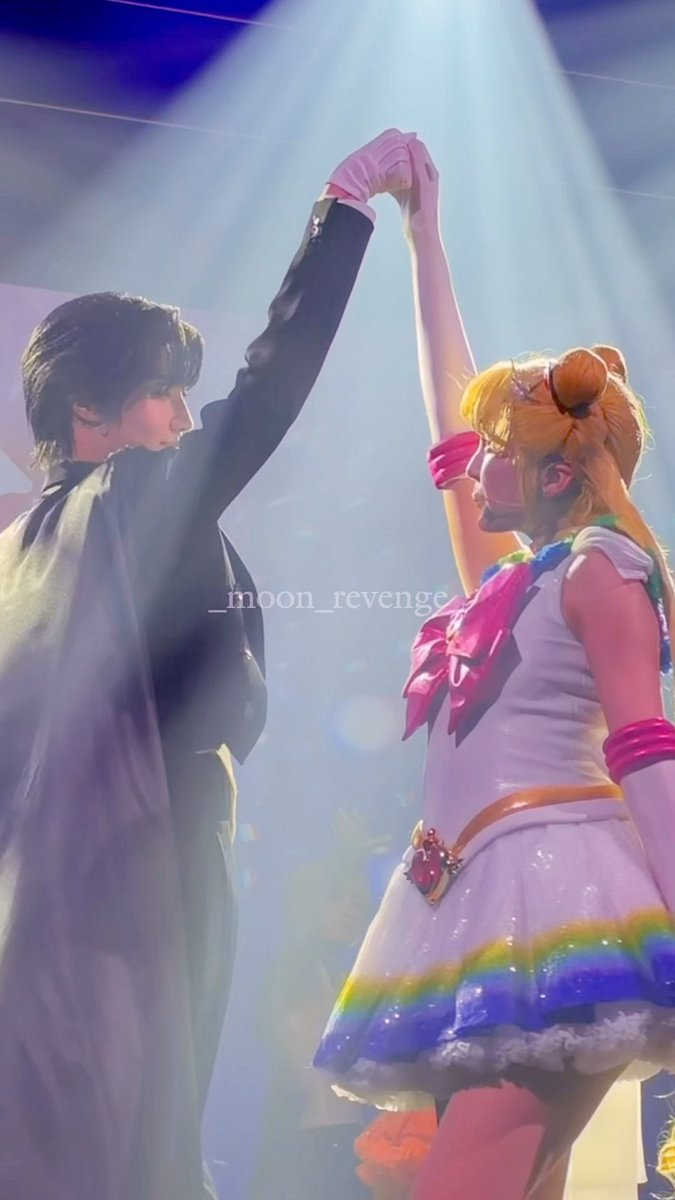 Two Sailor Moon cosplayers on stage strike a pose, raising their clasped hands together under bright spotlights, with the watermark '_moon_revenge_' visible.