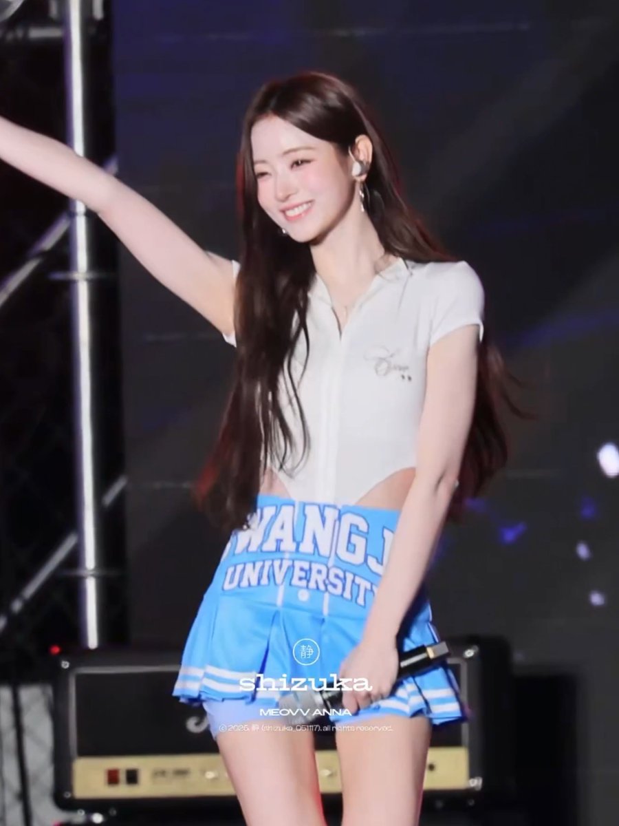 A woman wearing a white cropped top and a blue "SHANGHAI UNIVERSITY" cheerleader skirt smiles and waves on stage, with a watermark reading "静 shizuka" and "MEGV ANNA" visible across the skirt. Her cheerful pose and bright smile perfectly match the "so damn cute" sentiment of the post.
