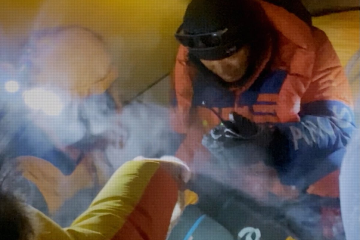 A rescuer in an orange and blue expedition jacket and black balaclava attends to an injured climber inside a yellow tent, with visible breath vapor suggesting extreme cold. The image captures the perilous conditions depicted in the documentary *The Last First Winter K2*, emphasizing the "high stakes and huge mistakes" referenced in the post.