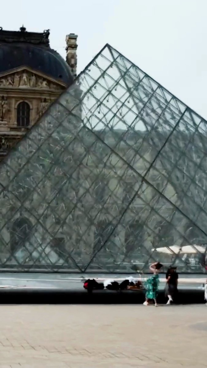 The glass Louvre Pyramid in Paris is shown on a sunny day, with people walking near its base.