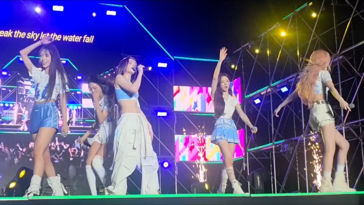 Five female performers are on stage at a concert with colorful lighting and screens, with on-screen text partially visible reading 'eak the sky let the water fall', as audience members hold up phones to record the performance.