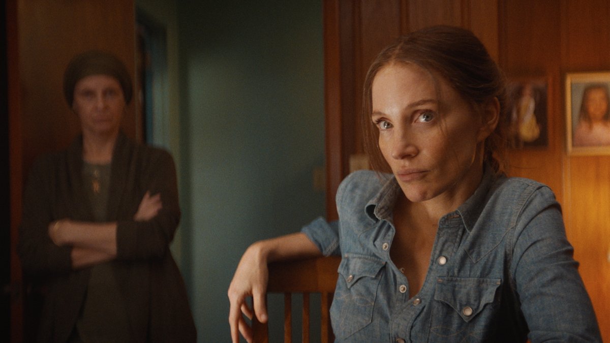 A woman with reddish-brown hair, wearing a denim shirt, sits in the foreground with an intense, contemplative expression, while another woman in a dark cardigan stands in the background with her arms crossed. The scene reflects the dramatic tone of the film "In the Heart of Nashville," premiering November 13 and starring Jessica Chastain, as it explores the emotional cost of fame in the music city.