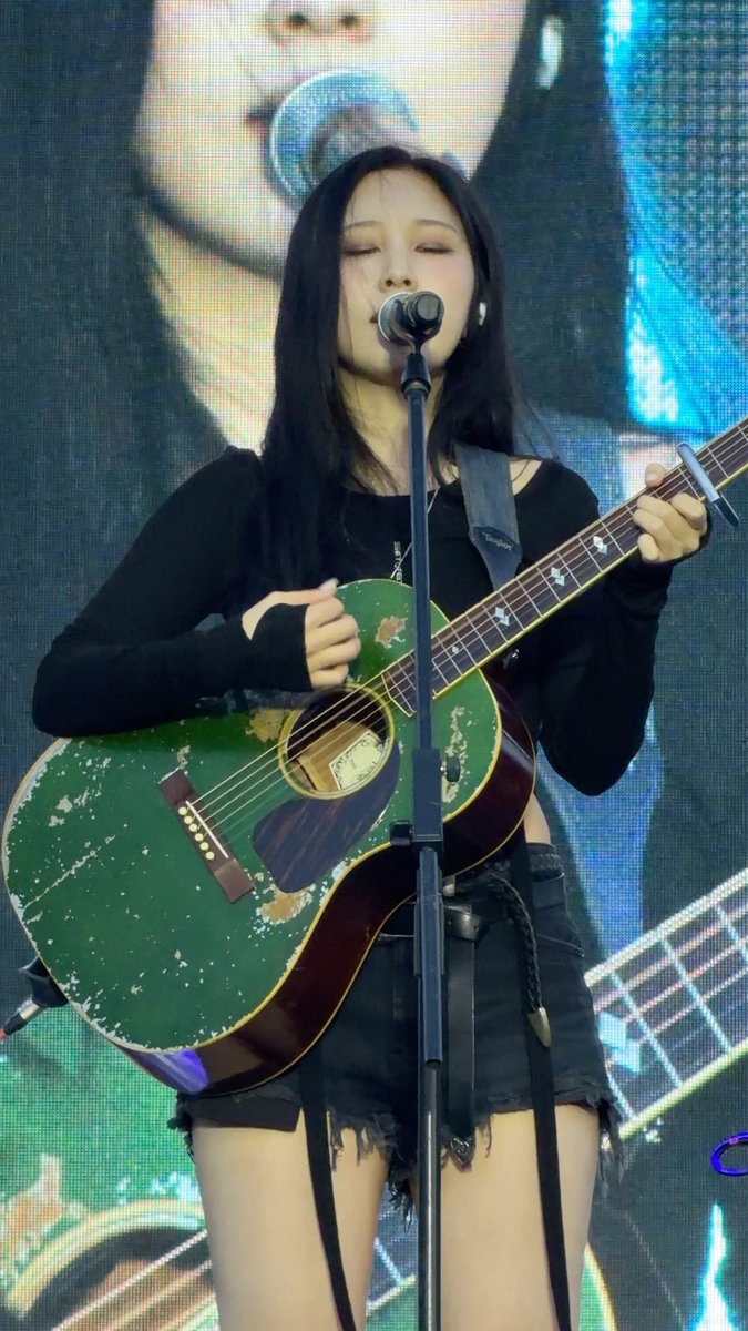 A woman with long black hair performs on stage, eyes closed, singing into a microphone while playing a weathered green acoustic guitar.