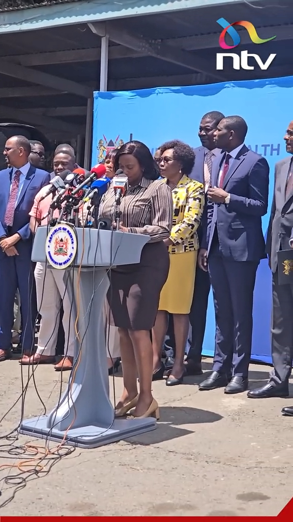 A woman in a striped blouse addresses reporters at a podium displaying the Kenyan government emblem, surrounded by officials during what appears to be an NTV Kenya press conference, following the country's first Ebola fatality in Nairobi.