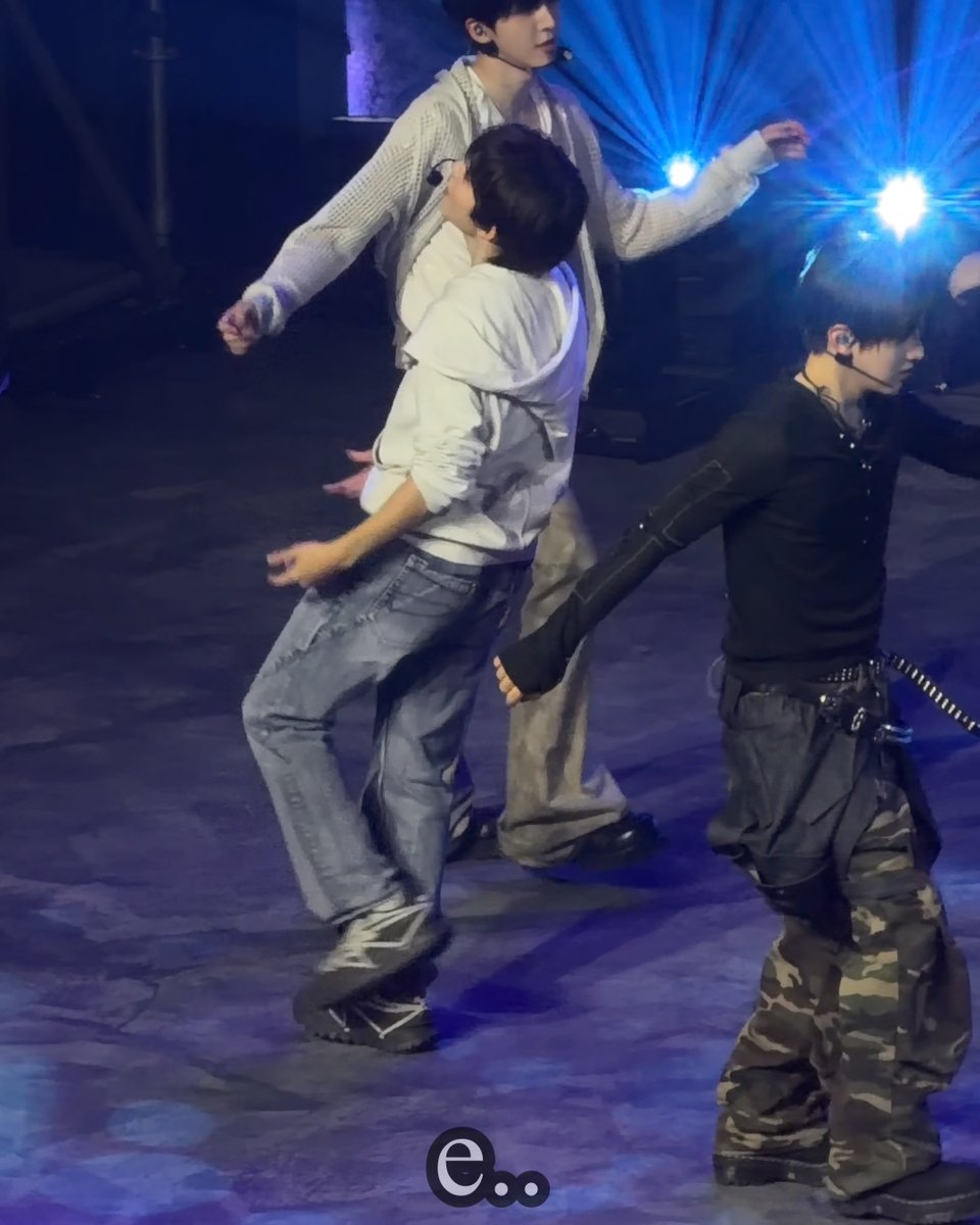 Three performers on a concert stage in dynamic poses; one wears a white graphic tee and baggy jeans, another a grey cardigan, and the third a black top with camo cargo pants.