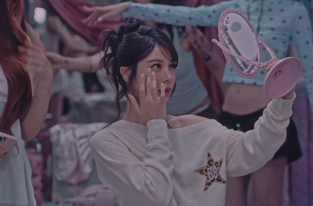 A young woman with dark hair in an updo wearing a white sweater with a star design holds a pink mirror while touching her face, seemingly backstage or in a dressing room area.
