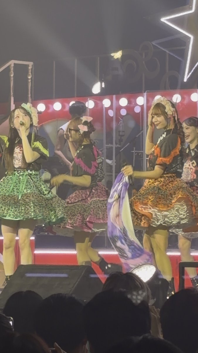 Members of a Japanese idol group performing on stage in colorful ruffled skirts and matching tops, with one singing into a microphone and another holding up a promotional poster.