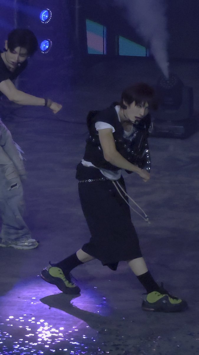 A concert photo showing two performers on stage bathed in blue light, with the main subject in the foreground wearing a black vest, shorts, and neon green and black sneakers while dancing.