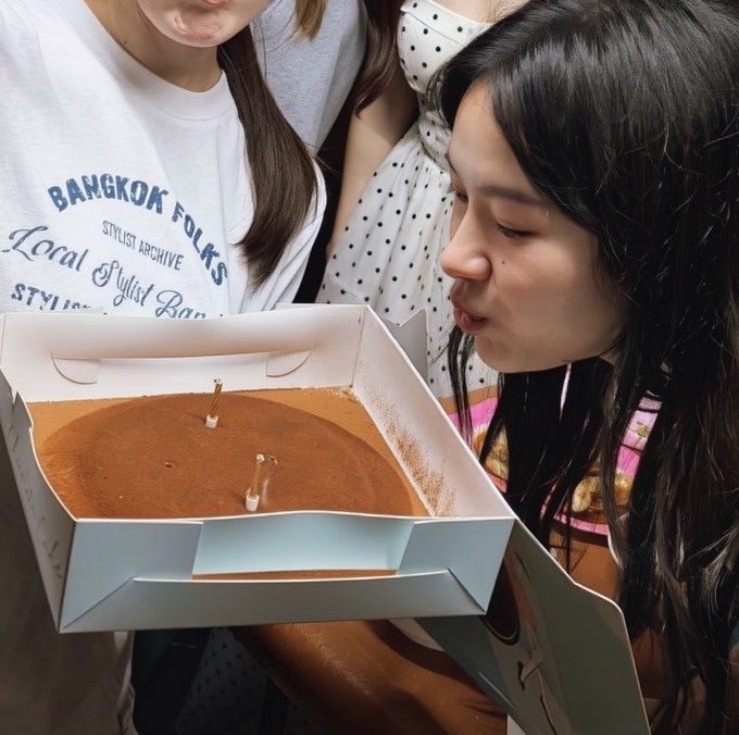 A woman blowing on a tiramisu cake in a box with lit candles, while others stand around her; one person wears a shirt reading 'BANGKOK FOLKS STYLIST ARCHIVE Local Stylist Boxer'.