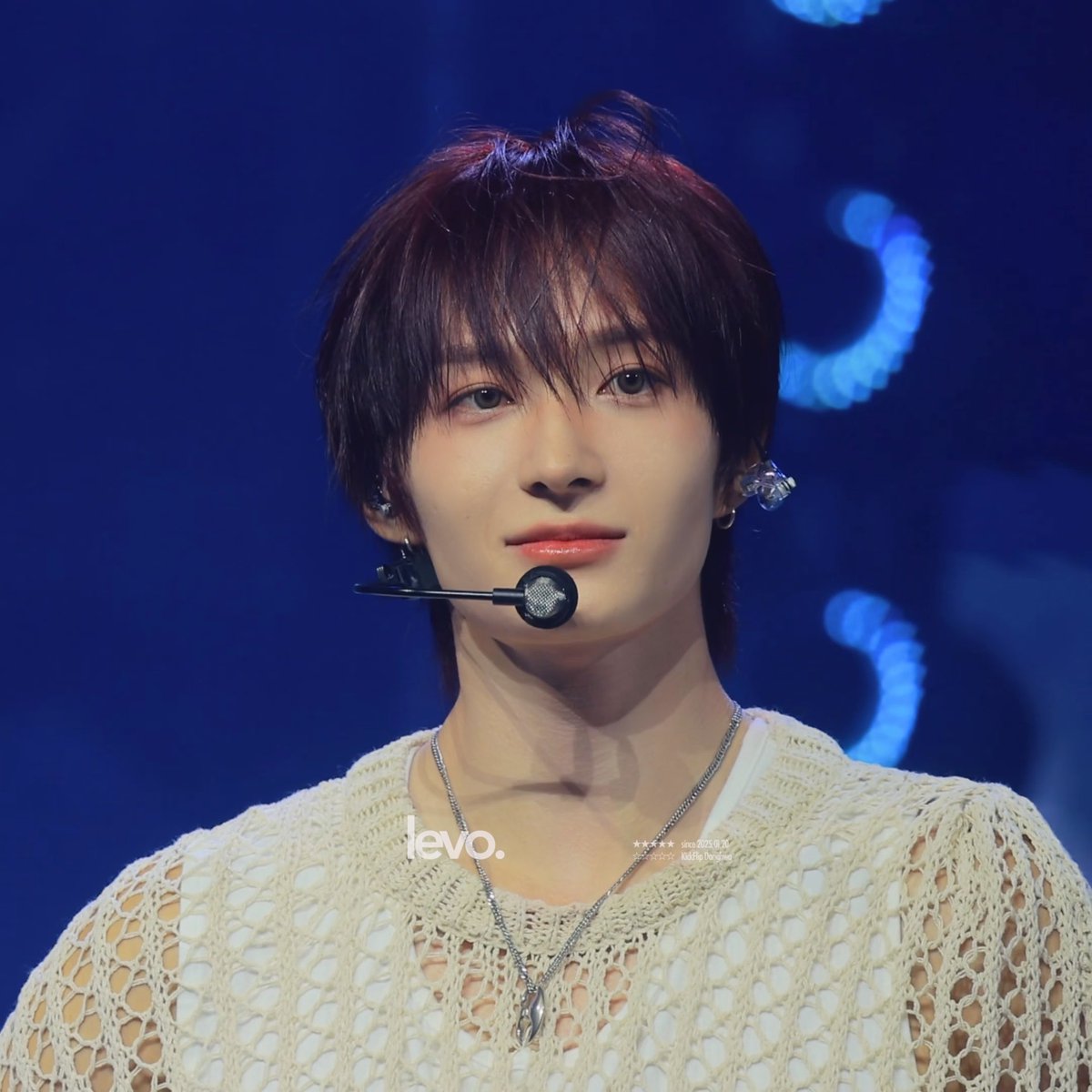 A close-up photo of a young male performer on stage wearing a cream-colored knit sweater, a headset microphone, and a silver pendant necklace, watermarked with "levo.".