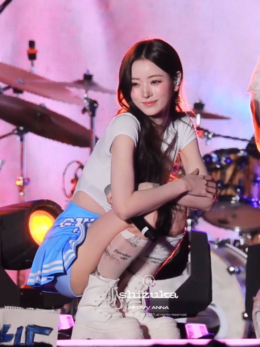 A performer crouches on stage wearing a white crop top and blue skirt, smiling at the viewer, with a watermark reading 'Shizuka / MEOVV ANNA' overlaid on the photo.