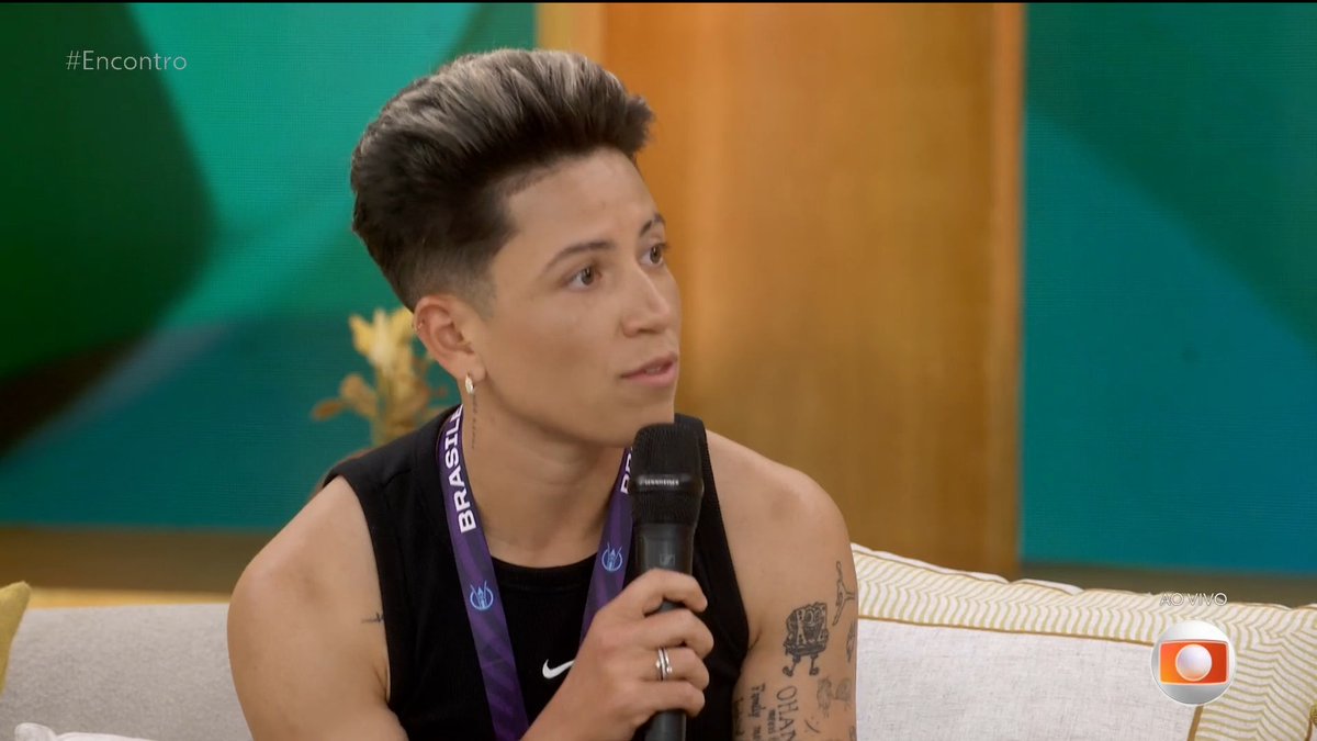 Person with short dark hair holding a microphone on the Brazilian TV show '#Encontro' (with Globo logo visible), wearing a lanyard that reads 'BRASÍLIA'.