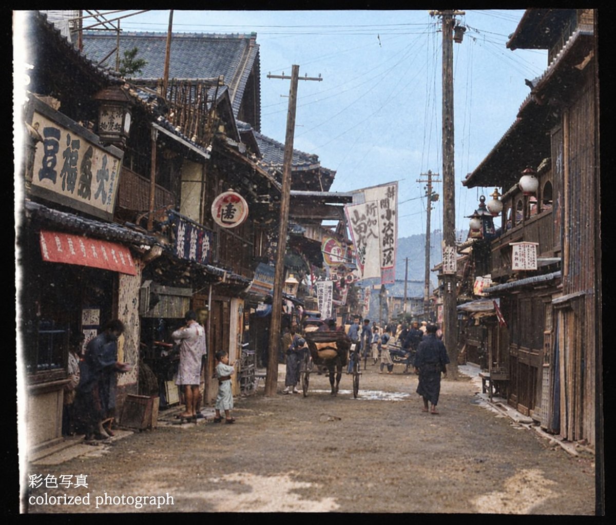 old_nippon's tweet photo. 1908年（明治41年）頃にアメリカの写真家によって撮影されたもの。伝統的な木造建築の商店が並ぶ、古い街並みが記録されています。
通りを行き交う人や人力車、当時の看板など、街の空気感が伝わってきます。
旗には「小次郎」や「新加入　山田」の文字が読めます。芝居小屋でしょうか。※彩色写真 https://t.co/uFGyI0PS55