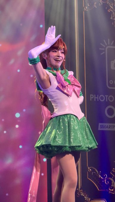 A woman wearing a Sailor Jupiter cosplay outfit from Sailor Moon, featuring a green skirt, white top with pink bow, and green choker, waves with a white-gloved hand on a stage.