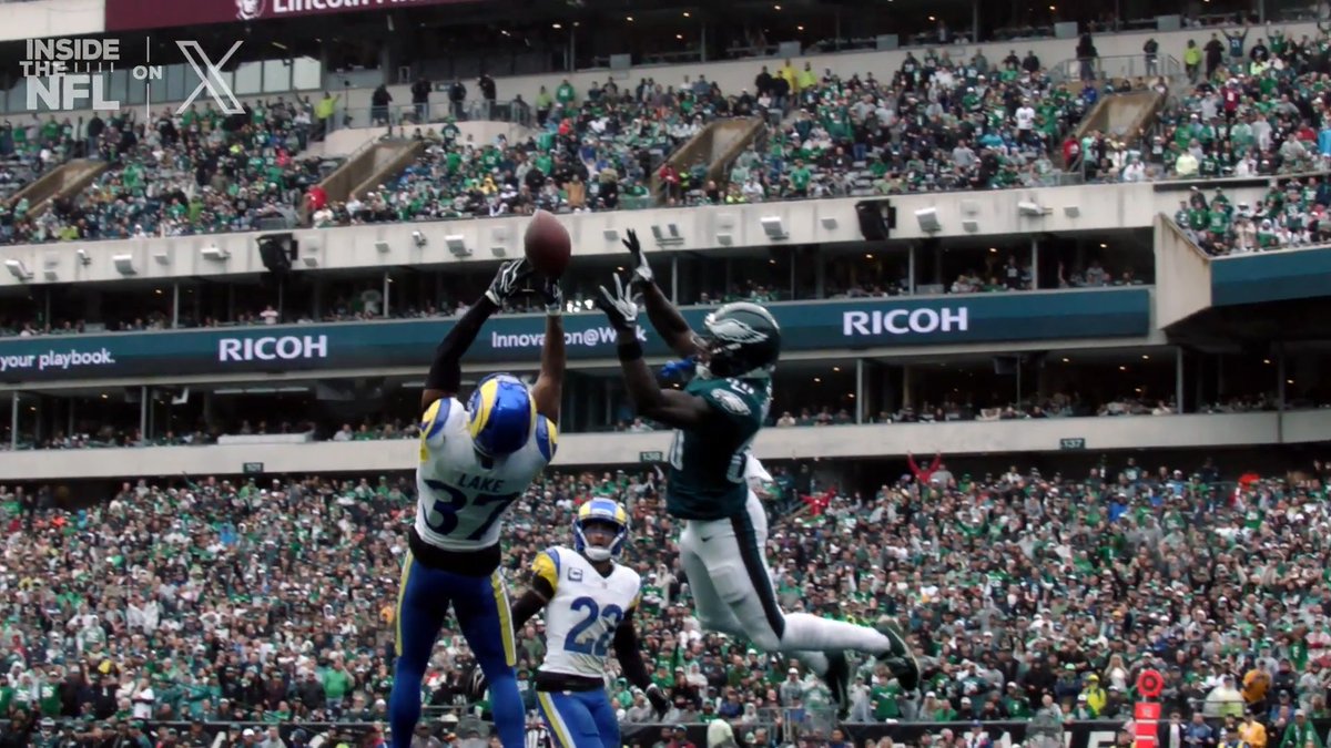 Two NFL players, #3 "Lake" in a Rams uniform and an Eagles defender, leap high into the air to contest a pass during a stadium game, with "RICOH" and "Innovation@Work" signage visible on the stadium walls behind them. The image captures the dramatic, slow-motion athleticism referenced in the post, showcasing the Eagles defender making a leaping play to break up the pass intended for the Rams receiver.