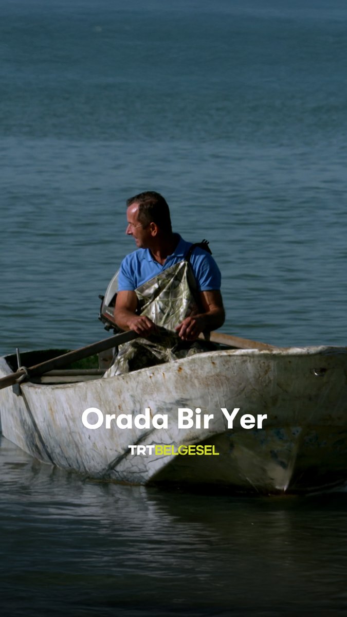 A man in a blue polo shirt rows a small white boat on calm water, holding a fish in a net, with the text 'Orada Bir Yer' and 'TRT BELGESEL' overlaid.