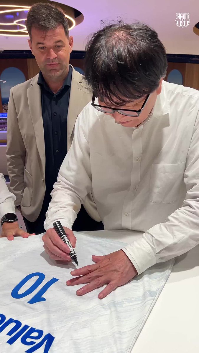 A man wearing glasses and a white shirt is signing a white FC Barcelona jersey with a black marker, while another man in a gray blazer stands nearby watching. The jersey features the letters "O" and "pien" visible in blue, and a FC Barcelona crest appears in the top right corner, consistent with the post about honoring Takahashi and his fictional Nankatsu SC team.