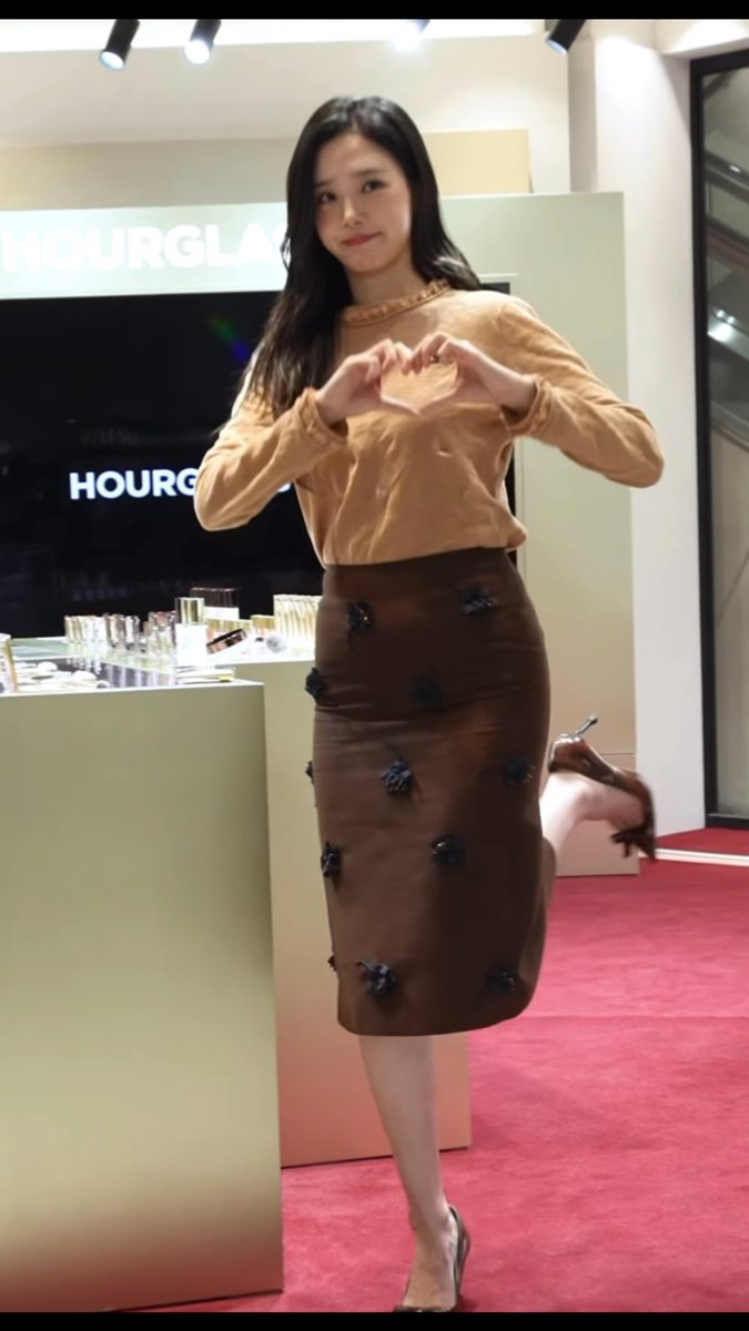 A woman in a tan blouse and brown skirt with black embellishments makes a heart shape with her hands at a HOURGLASS event on a red carpet.