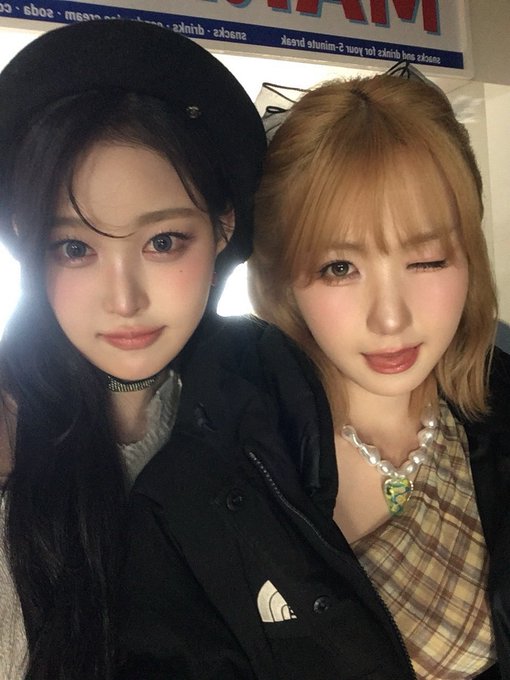 Two stylishly posed young women take a close-up selfie together, one in a black beret and dark jacket and the other with blonde hair wearing a pearl necklace with a green heart pendant and a plaid top, with partially shown text mentioning "snack - drinks - juice - beer -" and "Hiroshima" visible in the background. The image appears to celebrate a birthday for "Tommy Unnie" featuring hashtags related to the K-pop group Say My Name, including members #SOHA and #HITOMI.