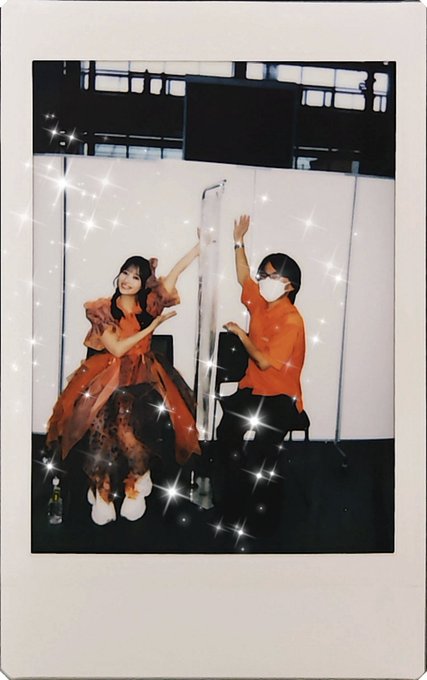A Polaroid-style photo shows a woman in an orange dress with a bow in her hair and a masked man in an orange shirt, both posing with raised arms in a room with sparkly filter effects.