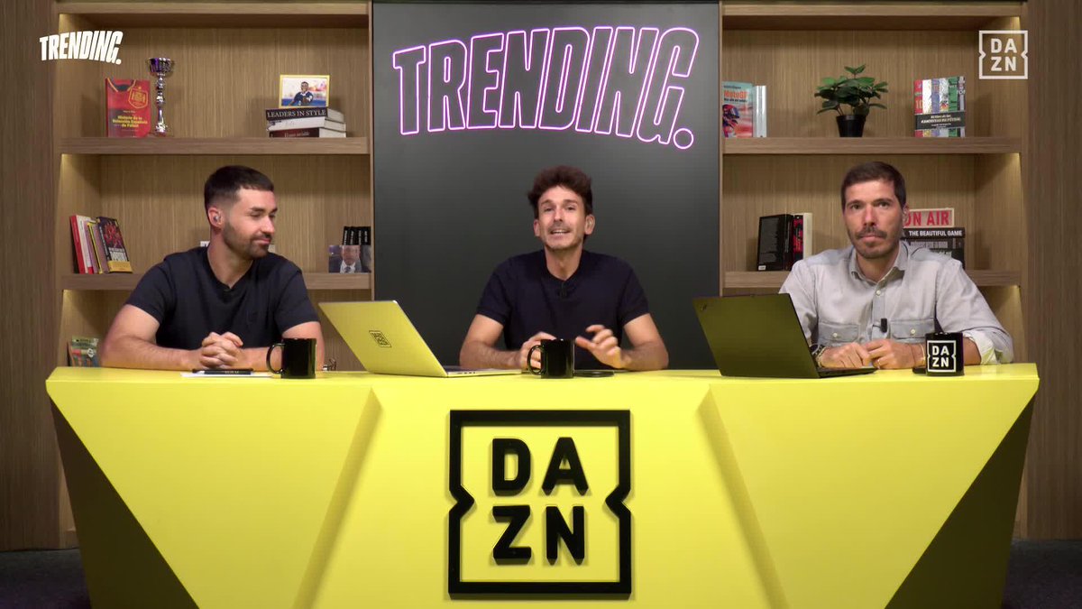 Three men sit at a bright yellow DAZN-branded desk with laptops and mugs, appearing on a show called "TRENDING" displayed in neon pink lettering behind them, likely providing commentary or analysis related to Mbappé's arrival at Valdebebas as referenced in the post.
