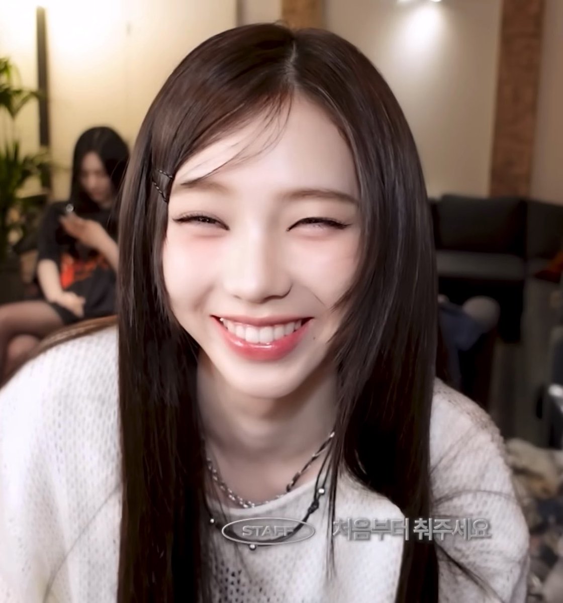 A close-up shot of a smiling young woman with long dark hair and hair clips, wearing a white knit top and layered necklaces.