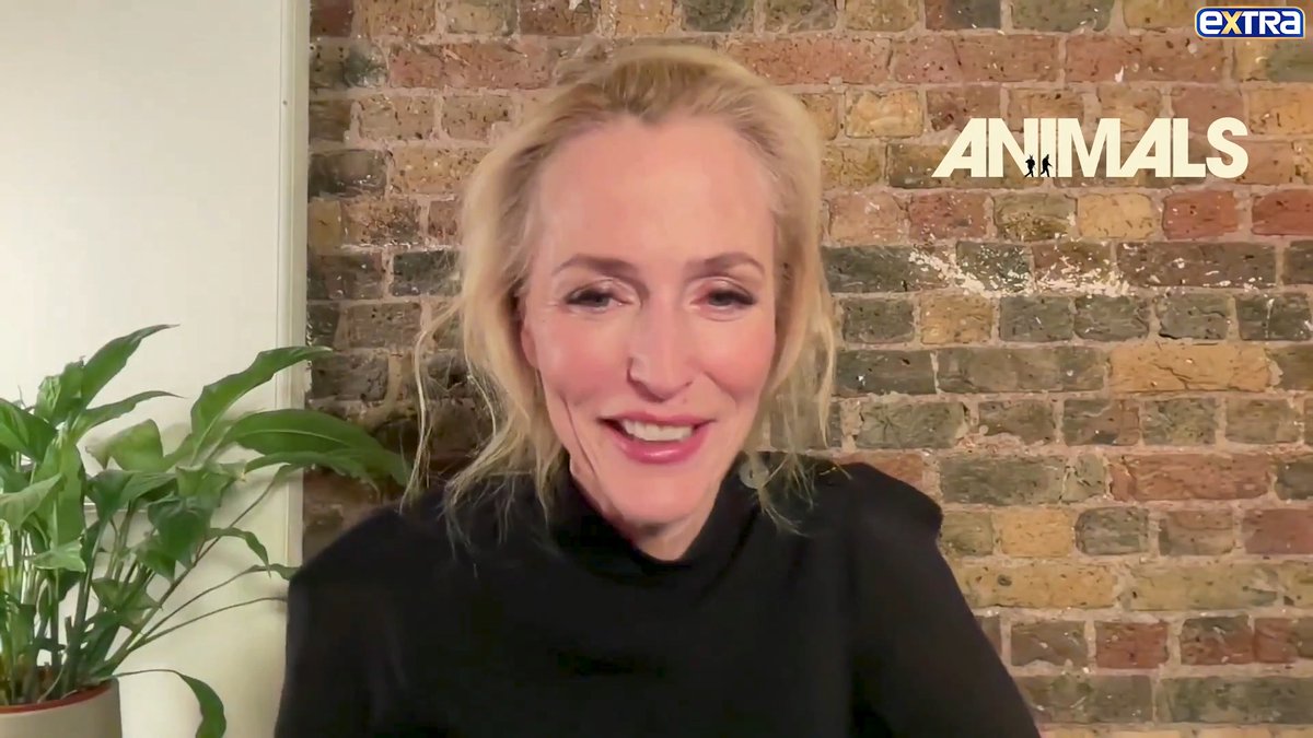 A blonde woman wearing a black turtleneck smiles during a video interview, with the 'EXTRA ANIMALS' logo visible in the upper right corner against a brick wall background.