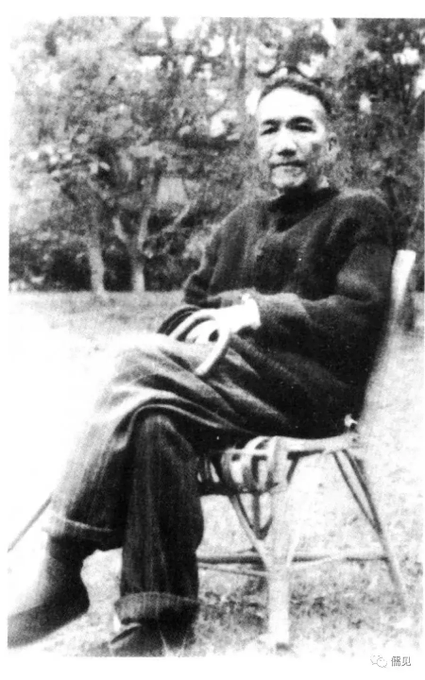 A vintage black-and-white photograph of an older man with short hair sitting relaxed in a white wicker chair outdoors, wearing dark traditional clothing and holding a book.