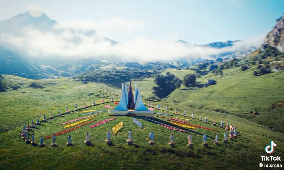 A scenic green mountain landscape featuring a decorative floral display and a spire-like structure in the center, with a TikTok watermark for m.srcha.