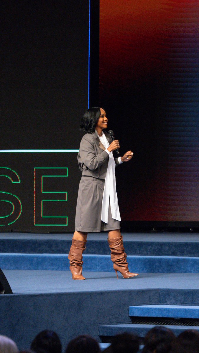 A woman with shoulder-length dark hair wearing a gray trench coat and brown slouchy boots speaks into a microphone on a stage with blue steps.