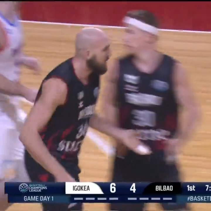 Two basketball players from Igokea (in dark jerseys) and Bilbao celebrate together on the court during Game Day 1 of the Basketball Champions League, with the scoreboard showing Igokea leading Bilbao 6-4 in the 1st quarter at 7:54.