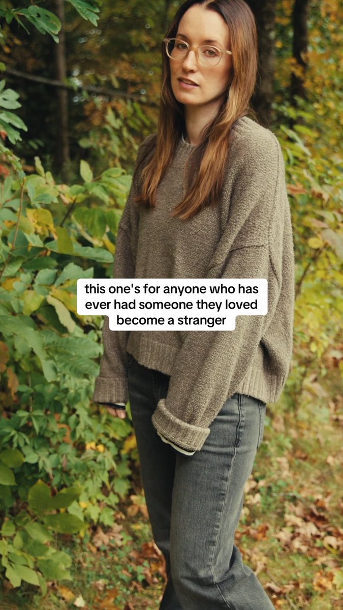 A woman with long brown hair and glasses wearing an oversized olive green sweater and dark jeans, standing in an autumn forest setting. A text overlay reads: 'this one's for anyone who has ever had someone they loved become a stranger'.