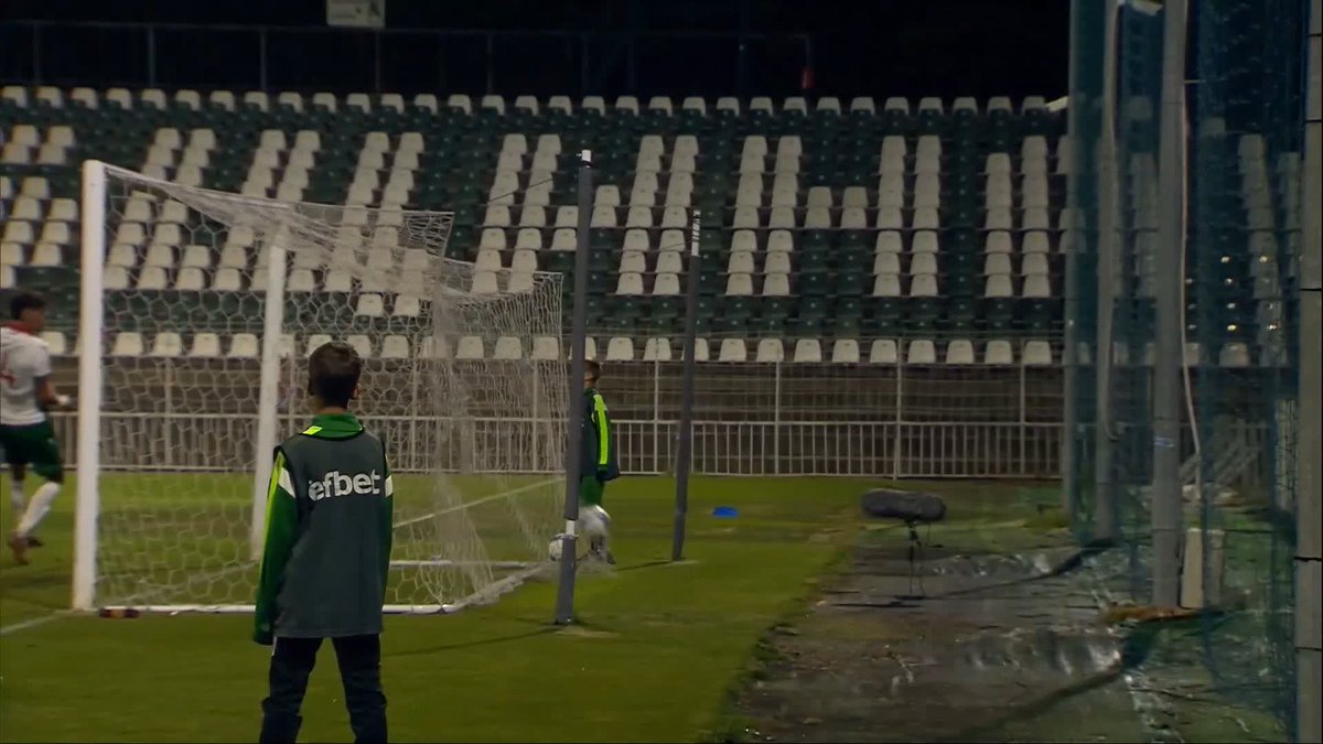 A young soccer player in a green "efbet" training bib stands on the pitch near the goal, with teammates and empty stadium seats visible in the background. The image captures a moment from a match in Bulgaria, fitting the caption of Bobby Wales scoring a second goal to seal victory in the #SCO21s #YoungTeam fixture.