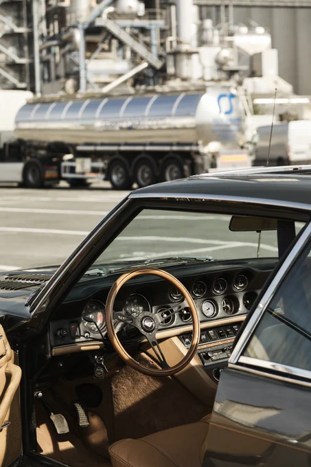The image shows the interior of a vintage car with a wooden steering wheel and classic dashboard controls. In the background, there's a tanker truck with a logo, parked near industrial equipment.