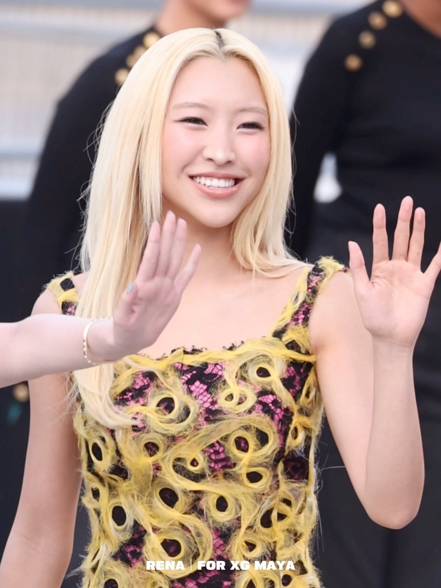 A blonde woman in a yellow and black patterned dress smiles and waves both hands, with text reading 'RENA | FOR XG MAYA' at the bottom.