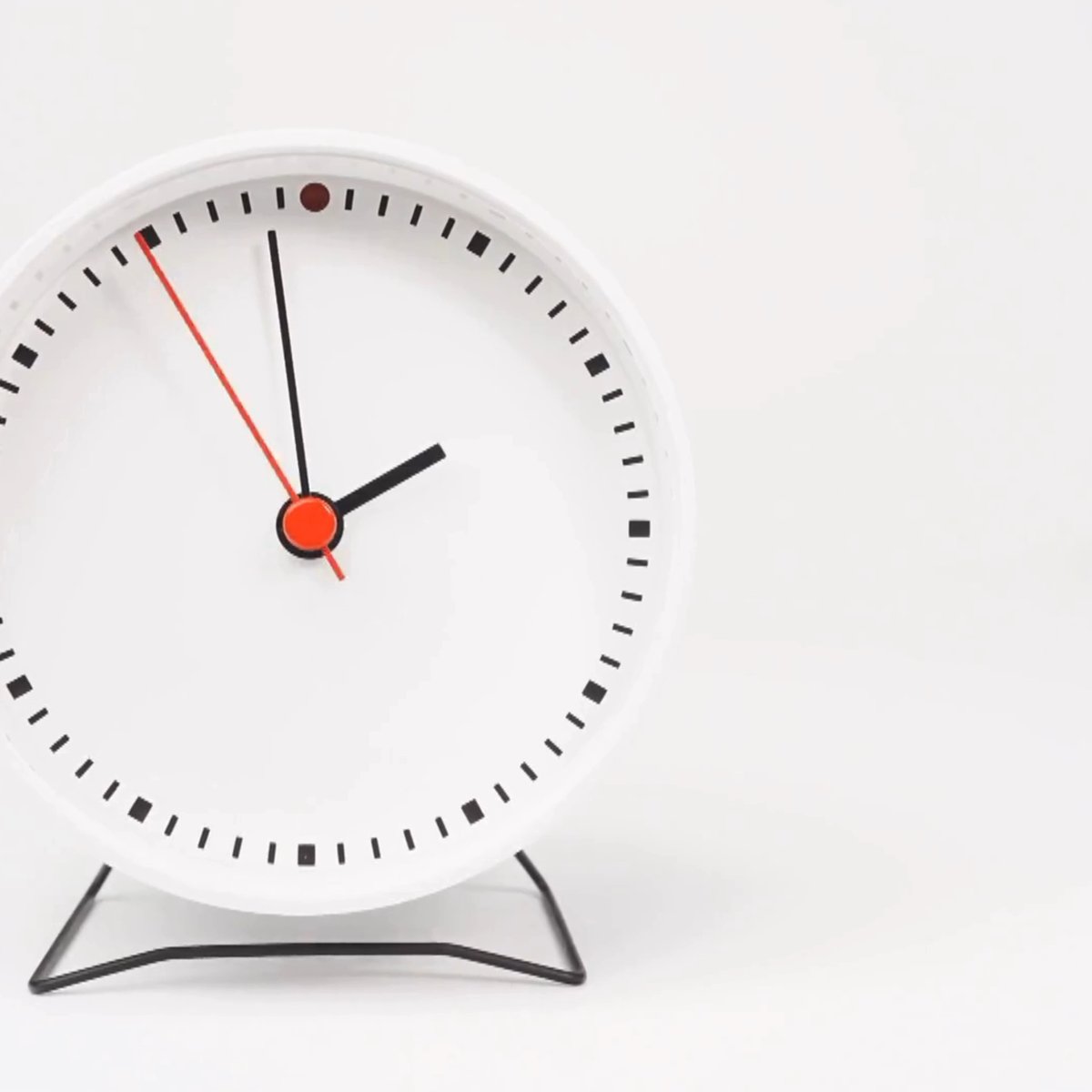 A minimalist white analog desk clock with black hour markers and hands plus a red second hand, photographed against a white background.