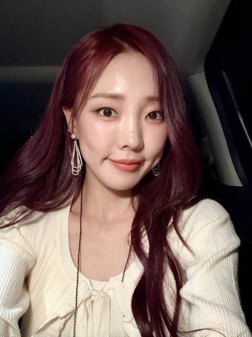 A selfie of a young woman with dark red hair wearing a cream-colored top and pearl earrings, sitting inside a car.
