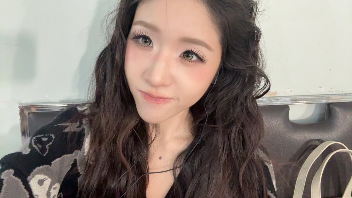 A close-up selfie of a young woman with long dark wavy hair, wearing dramatic makeup and a patterned sweater, sitting on a couch.