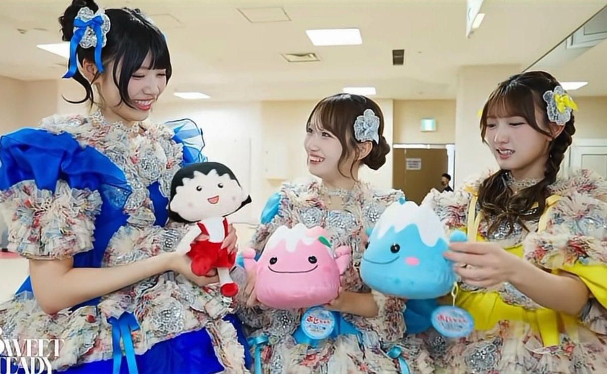 Three young women in elaborate floral-patterned dresses with blue, pink, and yellow accents, smiling while holding plush toys of a cartoon girl, a pink mountain character, and a blue mountain character.