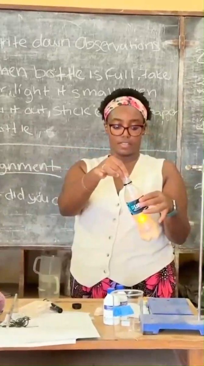 A still from an educational video showing a young woman standing in front of a chalkboard with partially visible text like "Write down observations" and "When bottle is full", holding a water bottle as part of a science experiment demonstration.