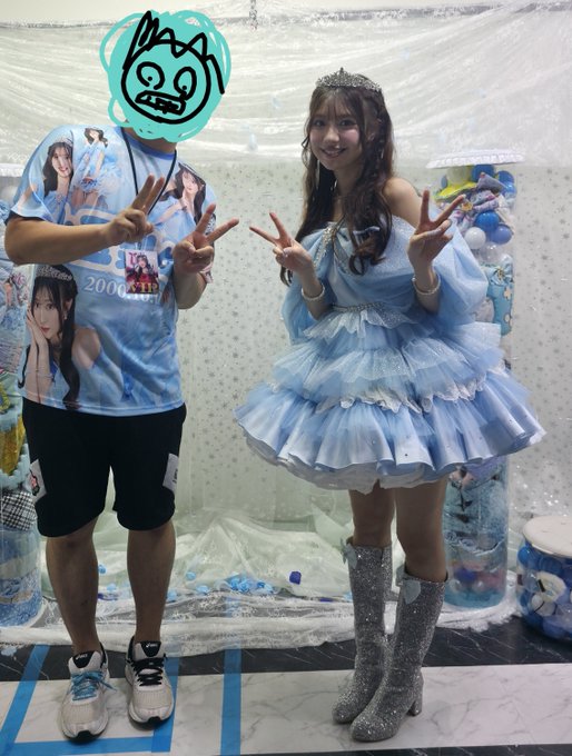 Two people posing with peace signs in front of a backdrop of packaged plush toys; one person's face is covered by a green doodle, and the woman wears a light blue princess dress.
