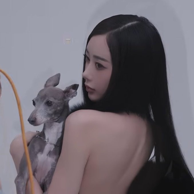A woman with long black hair holds a small greyhound-type dog over her shoulder, looking back toward the camera in a minimalist setting.