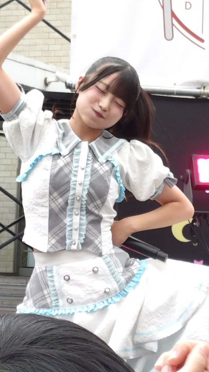 A young woman with dark hair in twin tails performs on stage in a white and gray plaid idol costume with light blue ruffled trim, holding her arm up with a microphone.